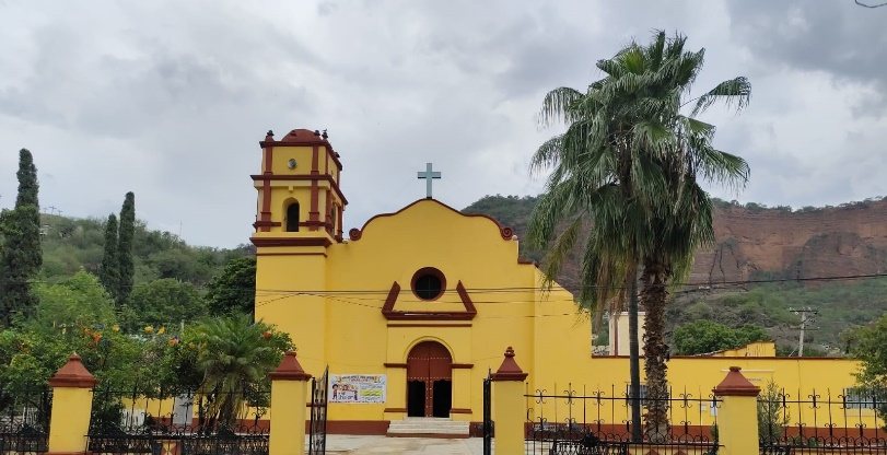 Templo de fachada amarilla con torre campanario y palmeras, frente a cerros rojizos