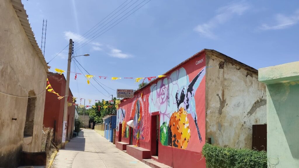 Calle con casas pintadas de rojo y un mural colorido, adornada con banderitas de papel