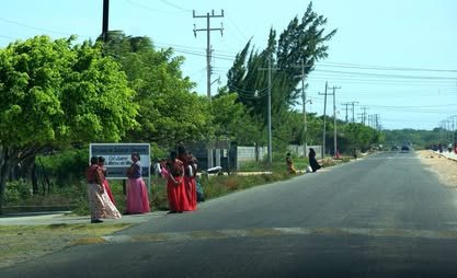 Carretera recta flanqueada por árboles; a un lado caminan mujeres con enaguas rojas