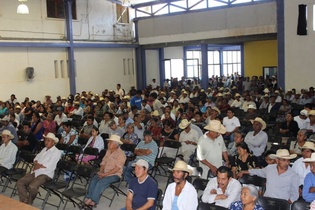 Memoria de campo 2011–2026 Salón amplio lleno de personas de la comunidad, muchas con sombrero, sentadas frente a una mesa durante una asamblea