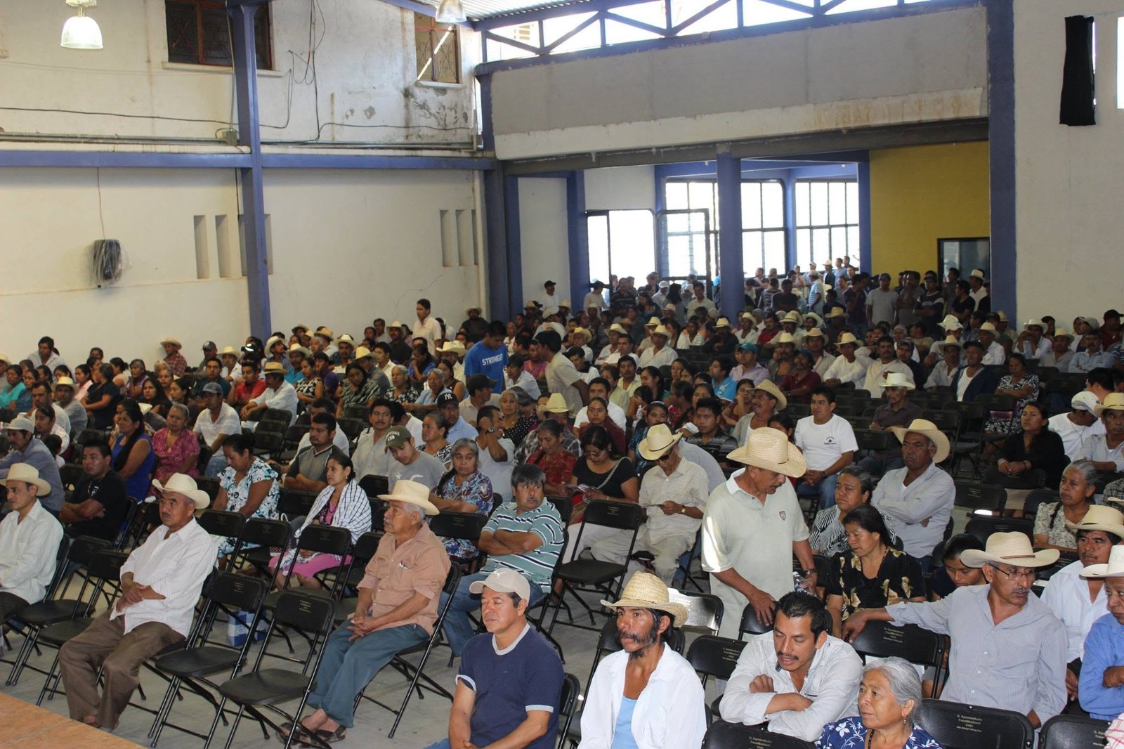Salón amplio lleno de personas de la comunidad, muchas con sombrero, sentadas frente a una mesa durante una asamblea