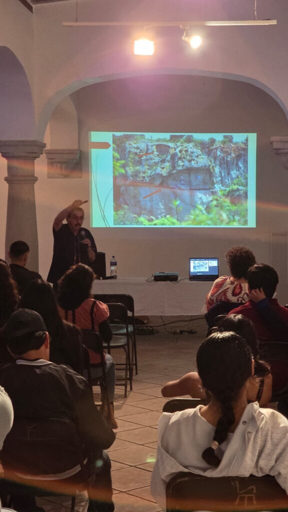 El Dr. Marcos Pedro Ramírez López, ponente en el conversatorio «El arte en Oaxaca a través del tiempo», en Mitla Asistentes, vistos de espaldas, siguen la presentación proyectada; al fondo, el ponente señala la pantalla