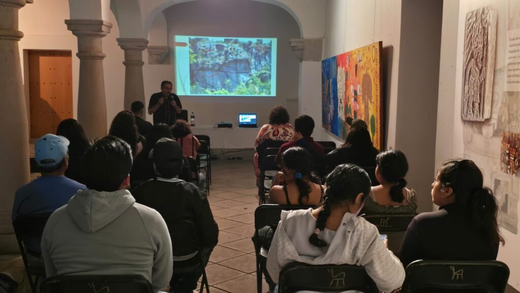 El Dr. Marcos Pedro Ramírez López, ponente en el conversatorio «El arte en Oaxaca a través del tiempo», en Mitla Sala del museo con público sentado de espaldas frente a la pantalla de proyección; a la derecha, un mural de colores