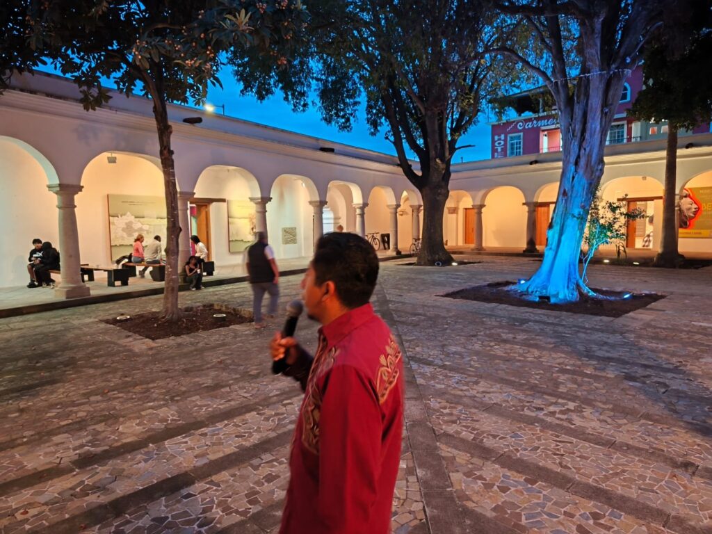 El Dr. Marcos Pedro Ramírez López, ponente en el conversatorio «El arte en Oaxaca a través del tiempo», en Mitla Patio porticado del museo al anochecer, con árboles iluminados; en primer plano, de espaldas, un presentador con micrófono