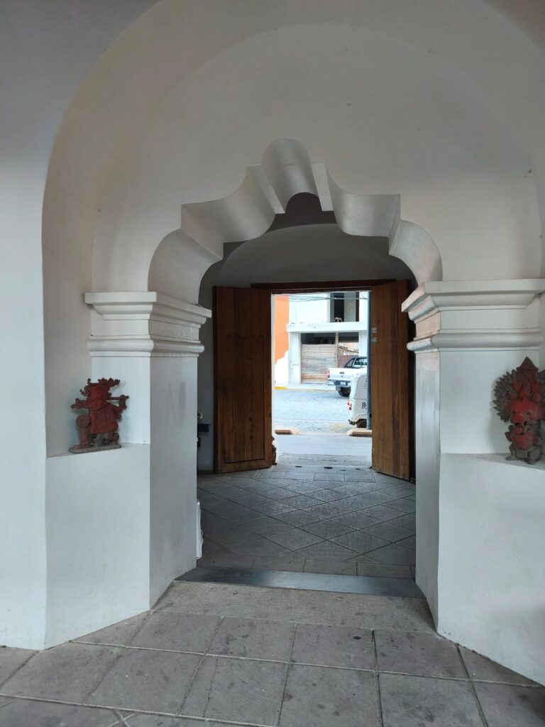 El Dr. Marcos Pedro Ramírez López, ponente en el conversatorio «El arte en Oaxaca a través del tiempo», en Mitla Acceso al museo: arco encalado con remate ondulado y portón de madera; a los lados, dos piezas de cerámica en nichos