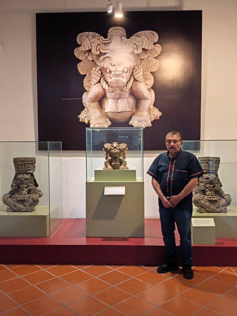 El Dr. Marcos Pedro Ramírez López, ponente en el conversatorio «El arte en Oaxaca a través del tiempo», en Mitla El Dr. Marcos Pedro Ramírez López de pie en una sala de exhibición, frente a urnas zapotecas en vitrinas