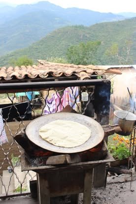 Tortilla sobre un comal de barro al aire libre, con techos de teja y montañas al fondo