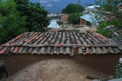 Techo de teja de barro de una casa, con árboles y casas al fondo