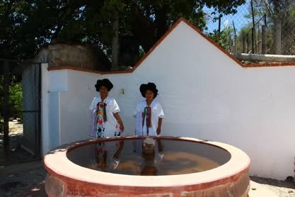 Fuente circular de piedra junto a un muro blanco; dos mujeres con traje tradicional blanco al fondo